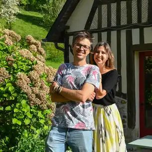 Ein Gastgeberpaar steht lächelnd vor einem Fachwerkhaus in der Normandie