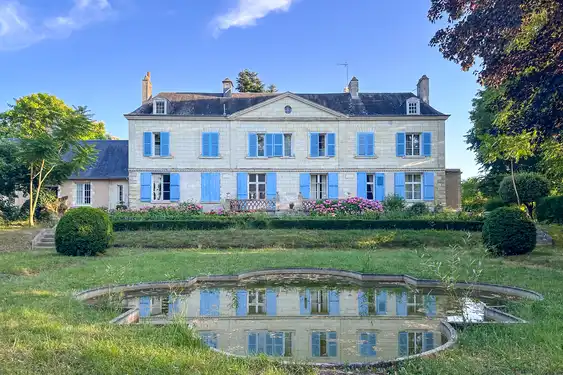 Ein Herrenhaus aus Tuffstein mit blauen Fensterläden und einem Teich vor dem Haus