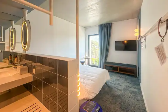 Ein Zimmer mit Waschbecken, dem Bett, dem gegenüber ein Fenster und daneben ein Fernseher