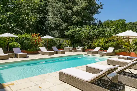 Großer Pool mit Rattanliegen und weißen Sonnenschirmen, umgeben von üppigem Grün und blühenden Sträuchern