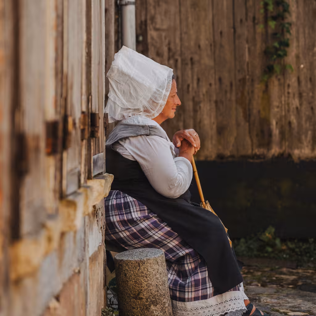 Eine Frau in bäuerlicher Kleidung sitzt vor einem rustikalen Haus in der Normandie